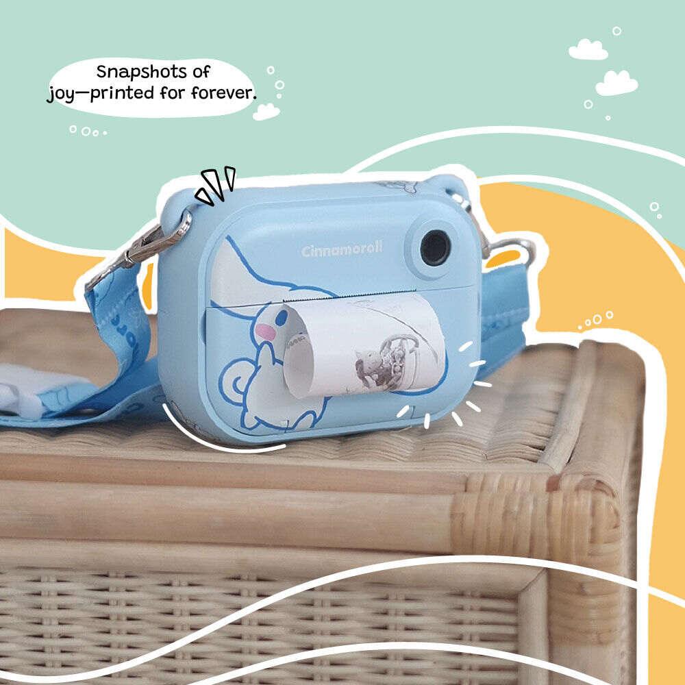 kiimento Model P Cinnamoroll kids’ digital camera placed on a wicker table, actively printing a black-and-white photo. The scene is framed by a colorful illustrated background with the caption “Snapshots of joy—printed for forever.”